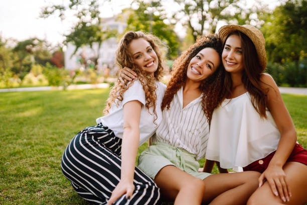 Trois femmes souriantes se tiennent ensemble dans un parc, symbolisant l'amitié et la joie de vivre. L'image met en avant l'unité et la force féminine dans un cadre naturel, avec des arbres en arrière-plan