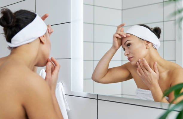 Une femme se tient devant un miroir en observant attentivement sa peau, mettant en lumière les soins qu'elle porte à son visage.